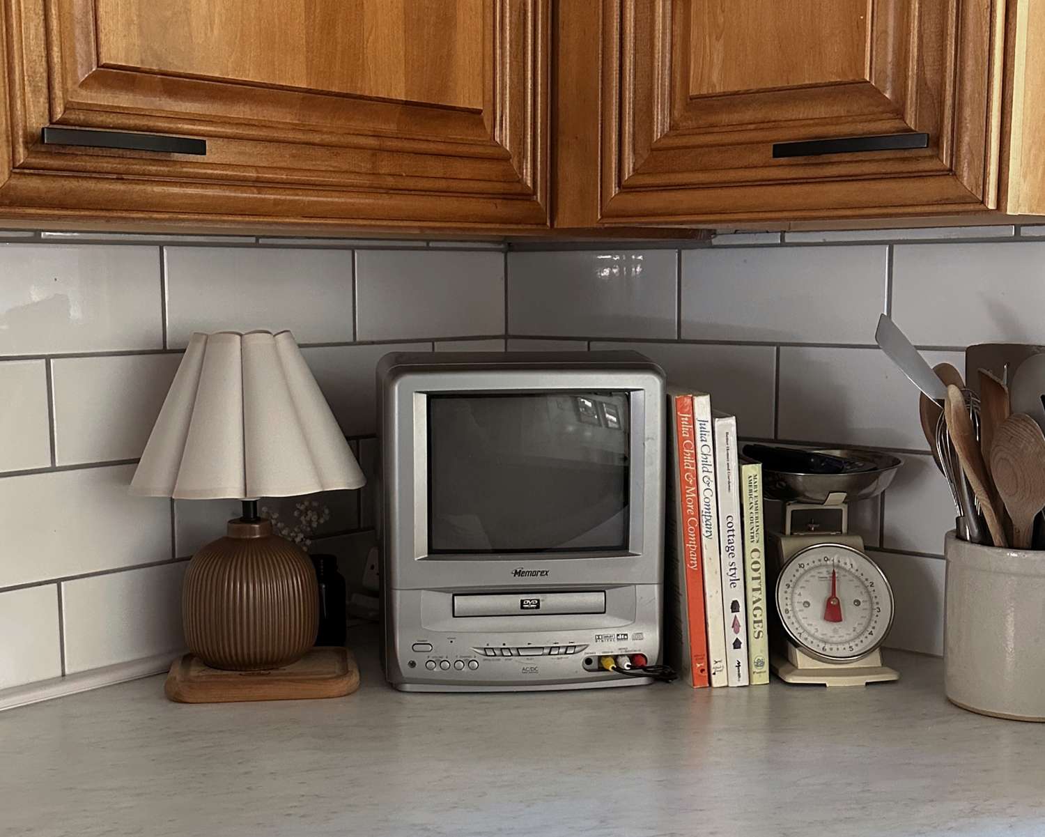 A small television sits on a kitchen counter surrounded by various items including books a lamp and a kitchen scale
