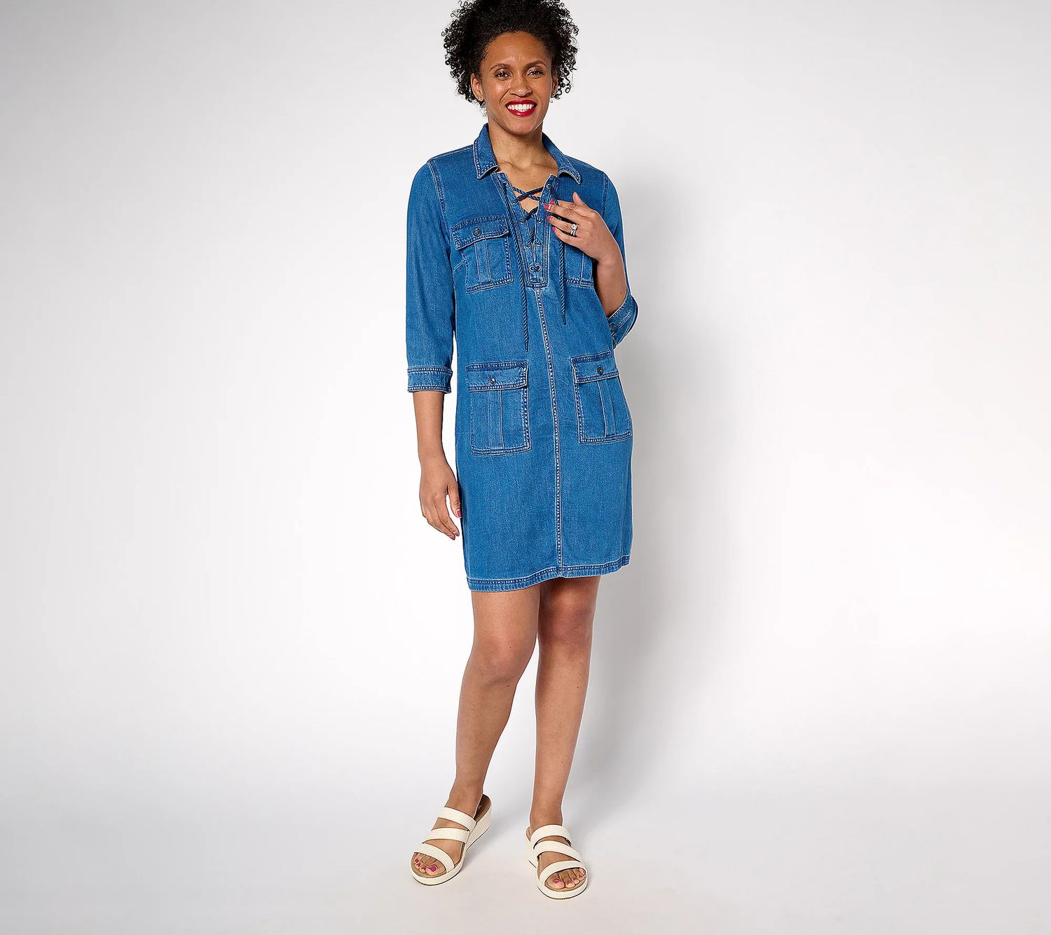 A smiling woman in a denim dress and sandals standing against a plain background. 