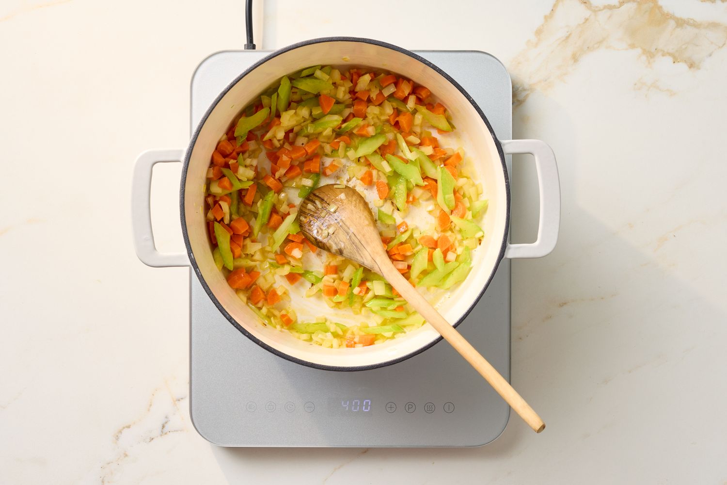 A pot on a stovetop containing sauted vegetables being stirred with a wooden spoon