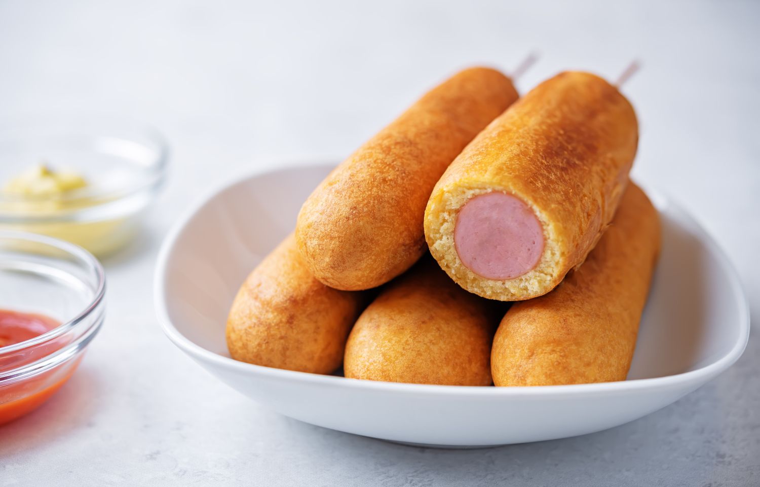 A white bowl containing several corn dogs one cut to show the inside