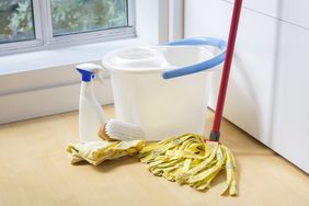 Cleaning tools including a mop bucket cleaning spray and cloths on a wooden floor near a window