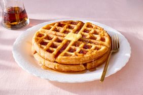 Two stacked waffles with a butter pat on a plate with a fork a drizzle of syrup and a syrup container in the background
