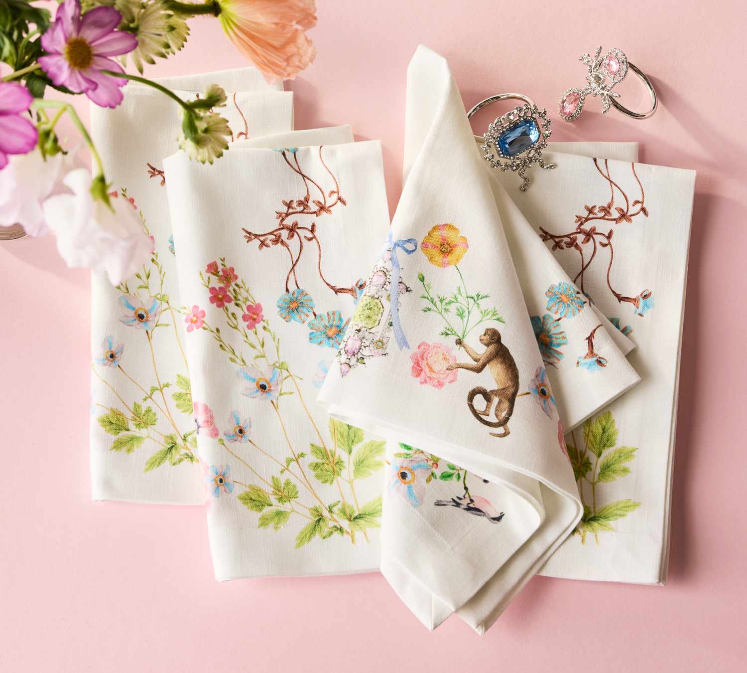 Floral-patterned napkins arranged with decorative jewelry on a light background, a flower bouquet in the corner