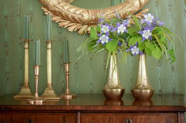 A wooden table with brass vases holding flowers and candlesticks a framed mirror on a greenpaneled wall in the background