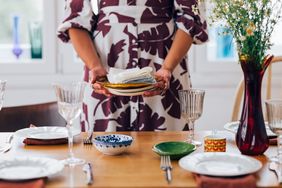 A person setting a dining table holding stacked plates with cloth napkins surrounded by dishes glasses and a vase of flowers