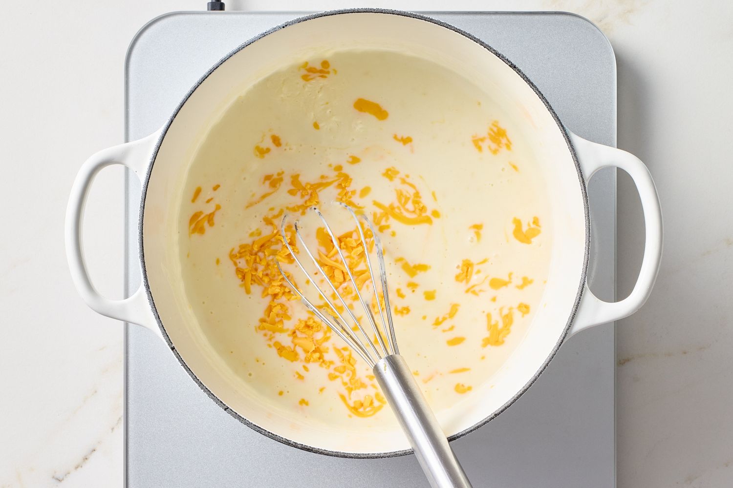 A cooking pot on a stovetop with a whisk and mixture that includes cheese