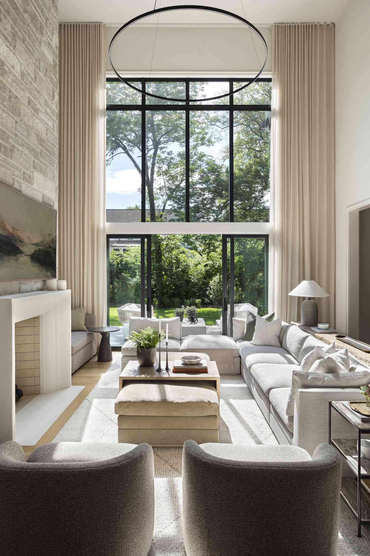 Minimalist living room by Brad Ramsey Interiors: white sectional sofa with textured pillows, stone fireplace, circular black chandelier, neutral rug, and large windows with garden views. Beige curtains, light wood floor, and natural light create a calm, stylish space.