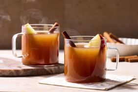 Two clear mugs of spiced cider with apple slices and cinnamon sticks steam rising