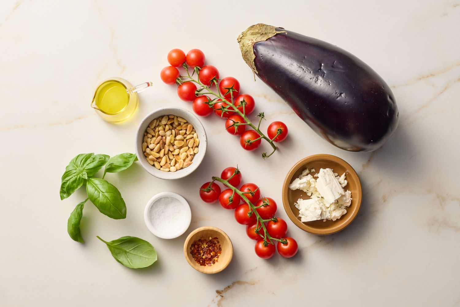 Ingredients for a dish including eggplant, cherry tomatoes, feta cheese, pine nuts, basil, olive oil, and seasoning