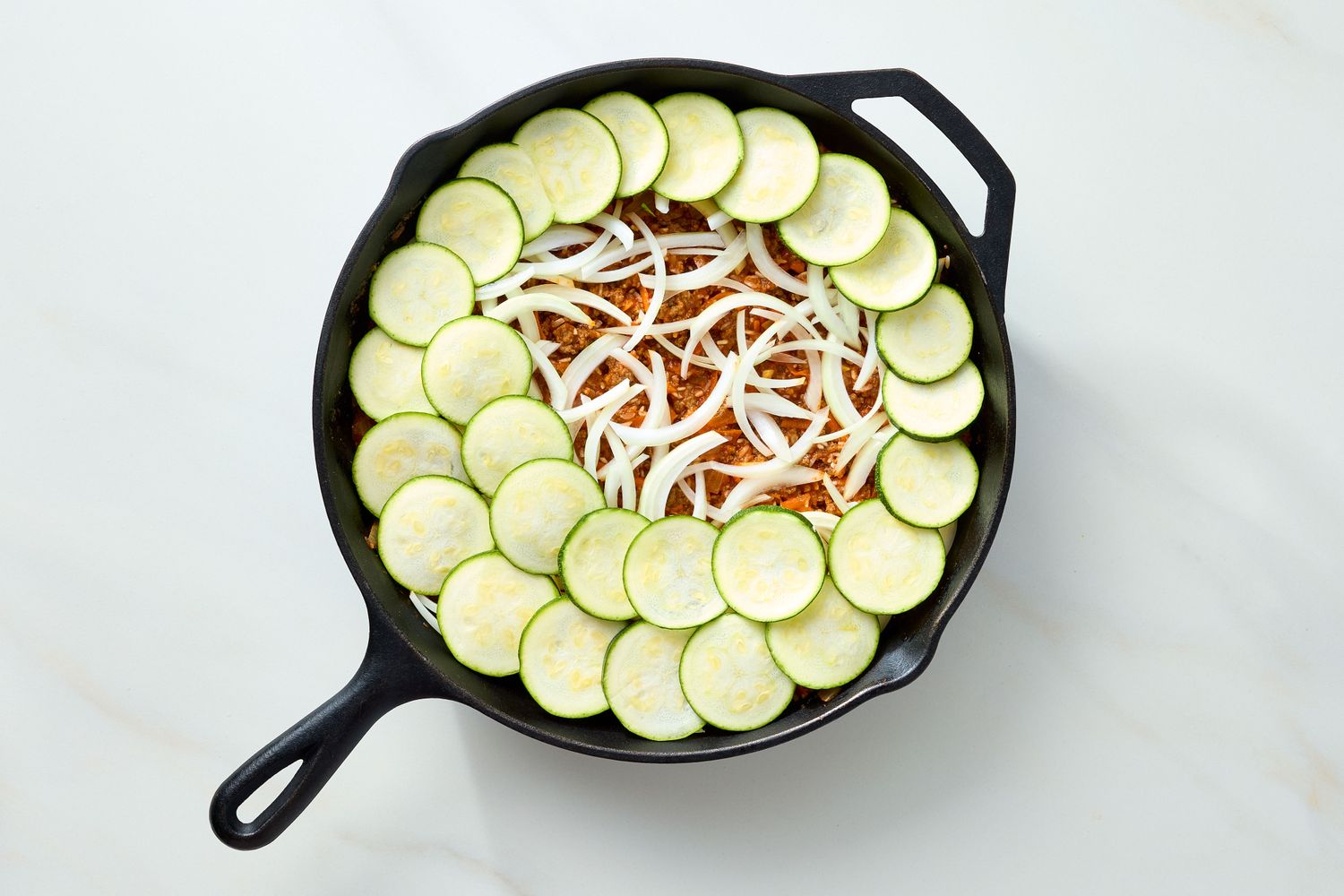 A casserole dish consisting of beef rice sliced zucchini and onions arranged in a round cast iron skillet