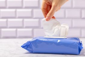 A hand pulling a wet wipe from a blue dispenser pack with a tiled background