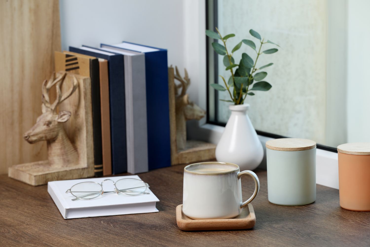 A wooden desk setup with books a coffee mug on a coaster eyeglasses on a notebook and a small plant in a vase