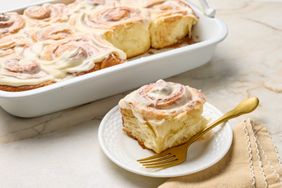 A serving of cinnamon roll on a plate with a fork with a tray of cinnamon rolls in the background