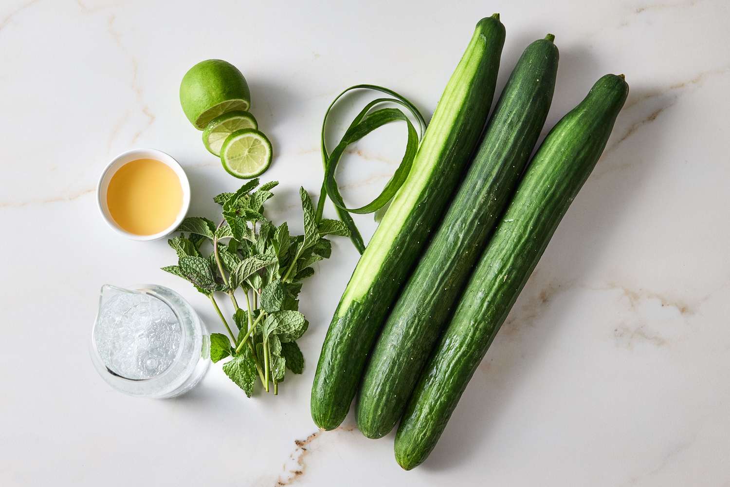 Ingredients for a cucumber cooler including cucumbers lime mint leaves and a glass of sparkling water on a marble surface