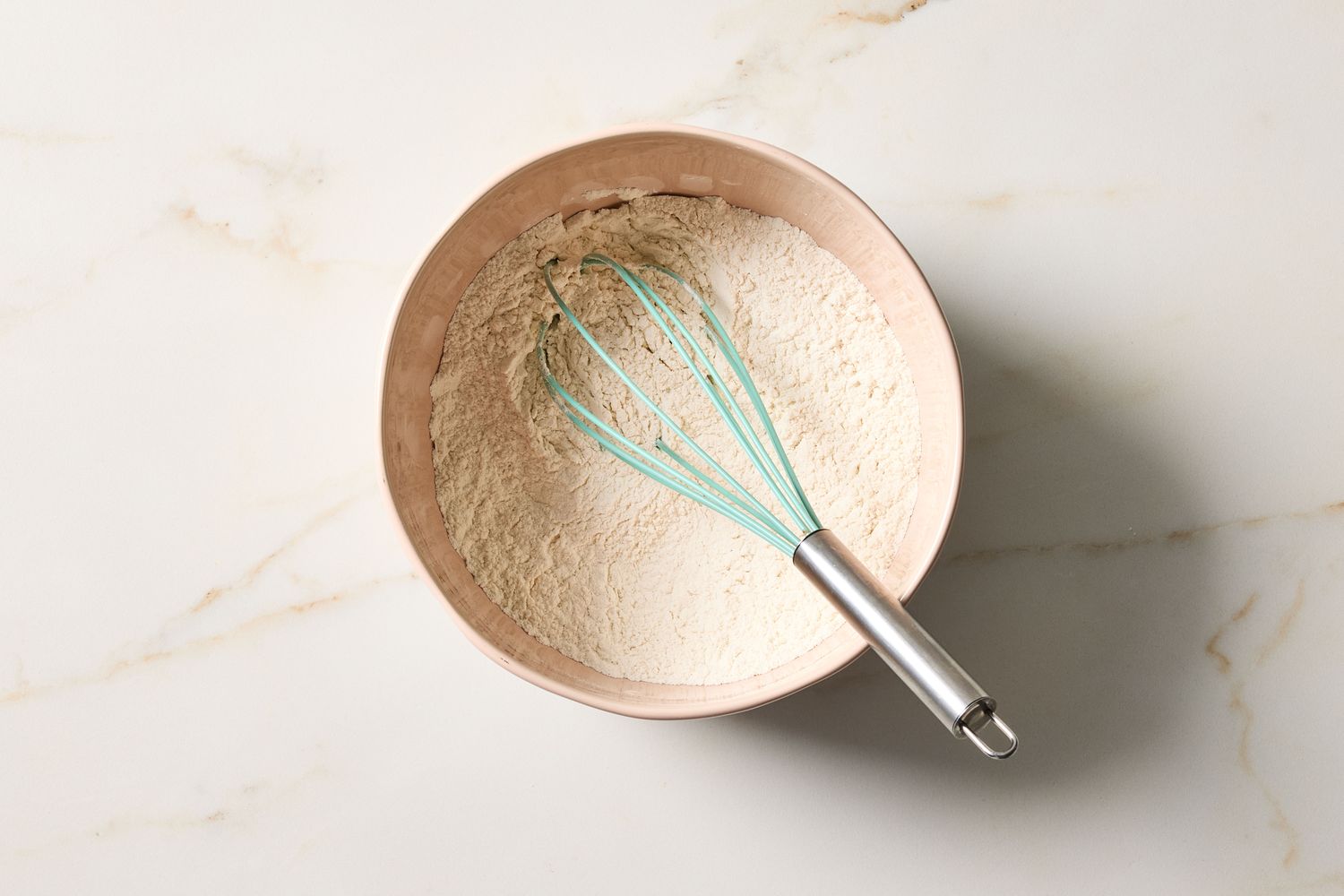 A whisk resting in a bowl of dry ingredients on a marble surface