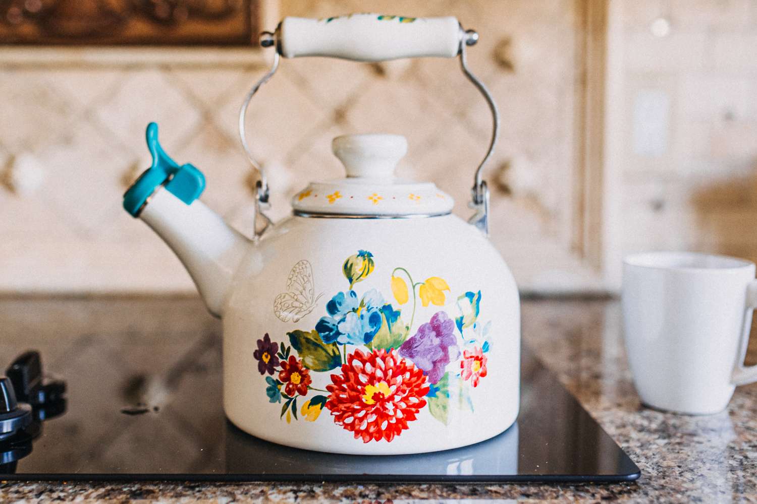 A decorative kettle with a floral design on a stovetop next to a white mug