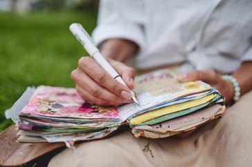A close-up shot of a person's hands writing in a notebook with a pen. The person is wearing a bracelet and a white shirt, and the background is blurred in beige tones. This image conveys a sense of focus and creativity.