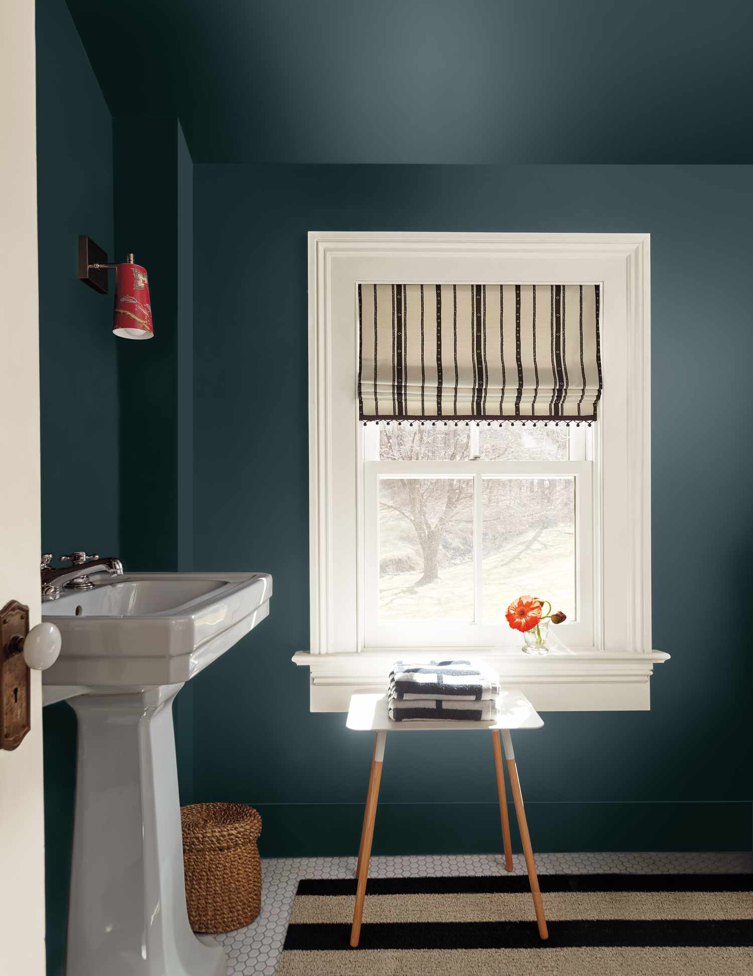 A bathroom with a pedestal sink a striped rug a small table with books and a flower vase near a window with a striped shade