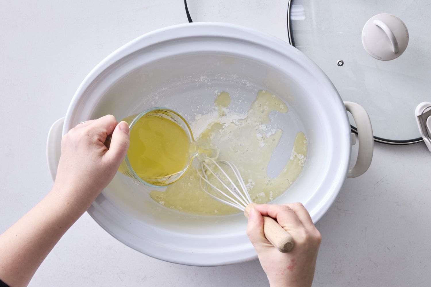 Hands mixing ingredients in a slow cooker with a whisk and pouring liquid from a cup