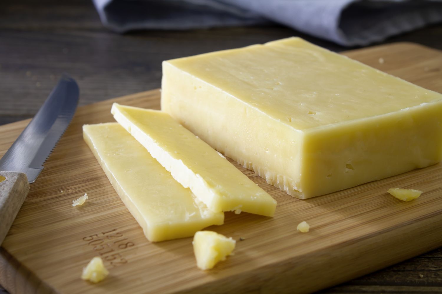 A block of cheddar cheese with some slices cut on a wooden cutting board a knife beside it