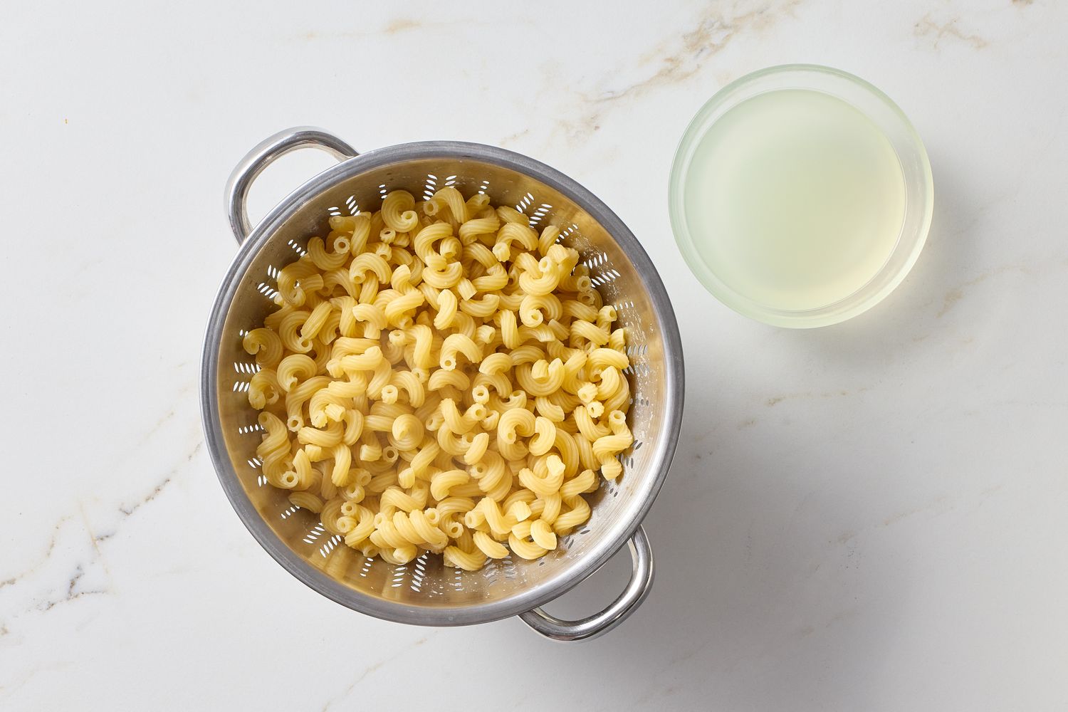 Cooked macaroni in a colander with a bowl of pasta water beside it