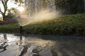 Sprinkler spraying water over a grassy lawn near a paved surface at sunrise creating a misty effect