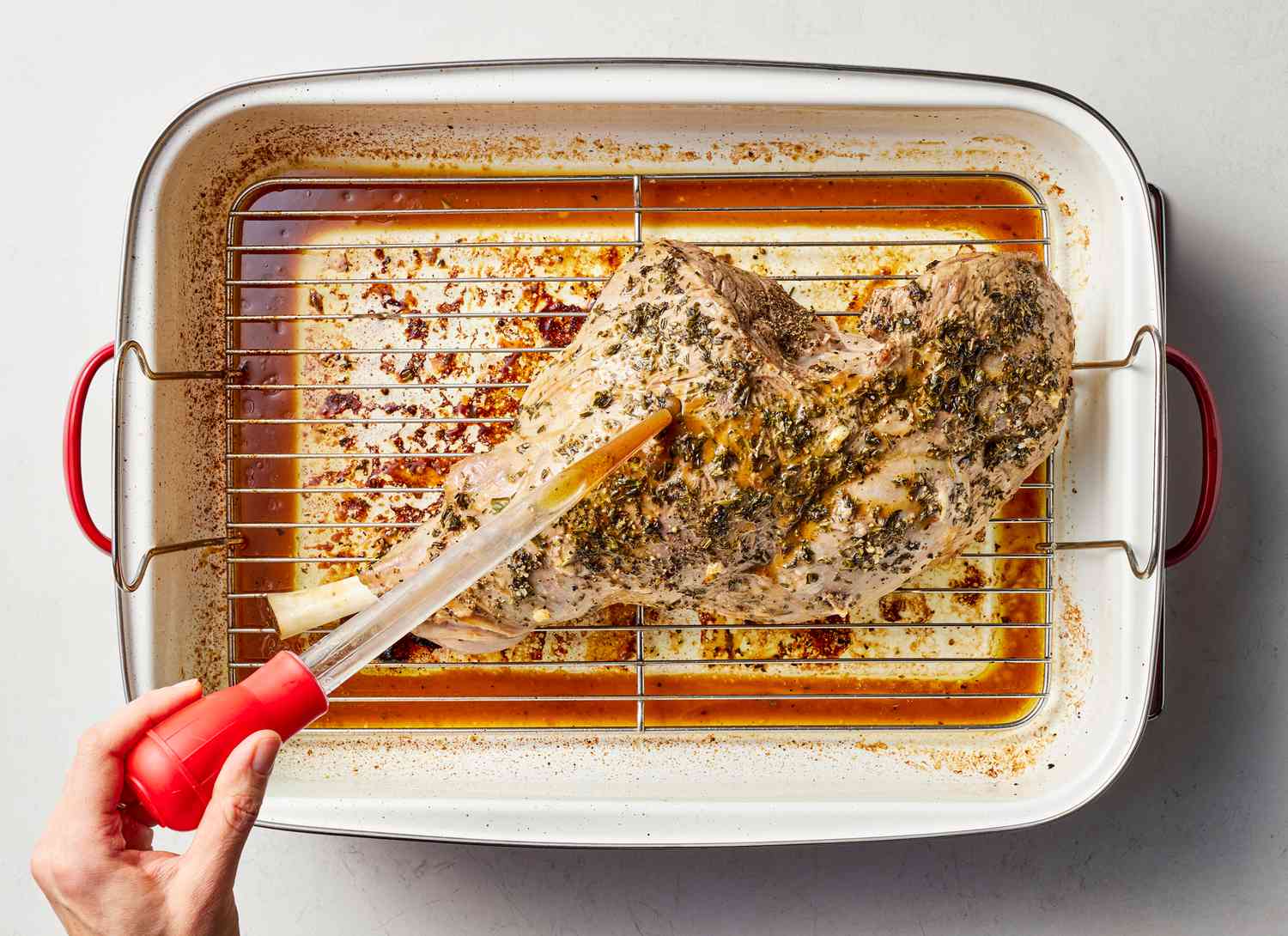 someone basting a leg of lamb in a roasting pan