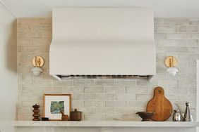 A kitchen with a range hood brick backsplash floating shelf holding decor and brass wall sconces