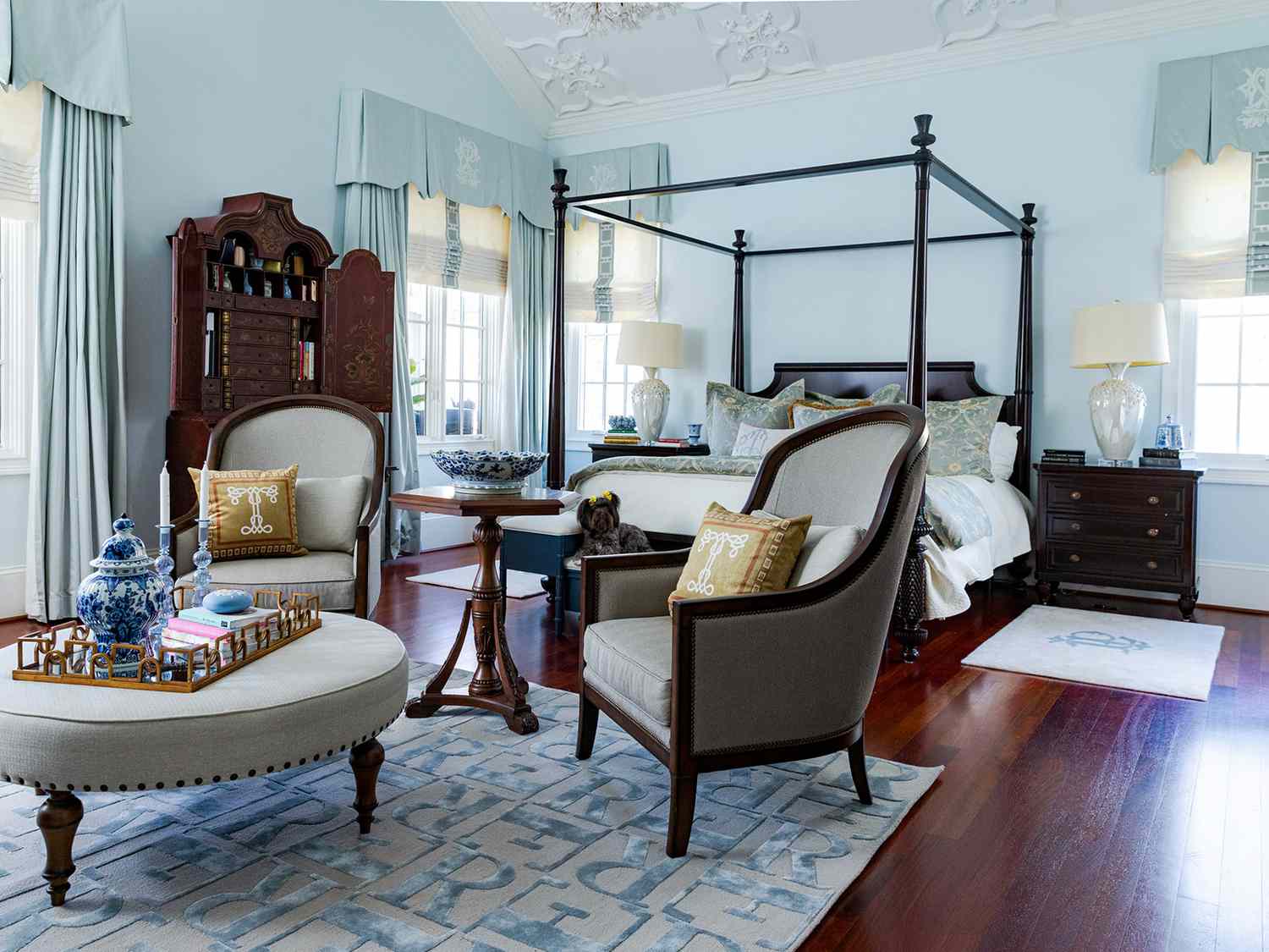 A pale blue Georgian style bedroom with antique wooden furniture and a graphic, initial rug in the foreground under a seating area.