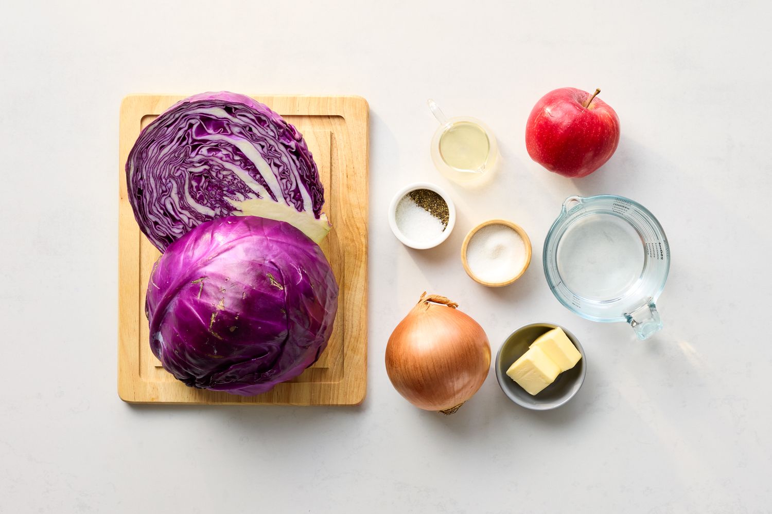 Ingredients arrangement showing red cabbage apple onion butter and seasonings on a cutting board and white surface