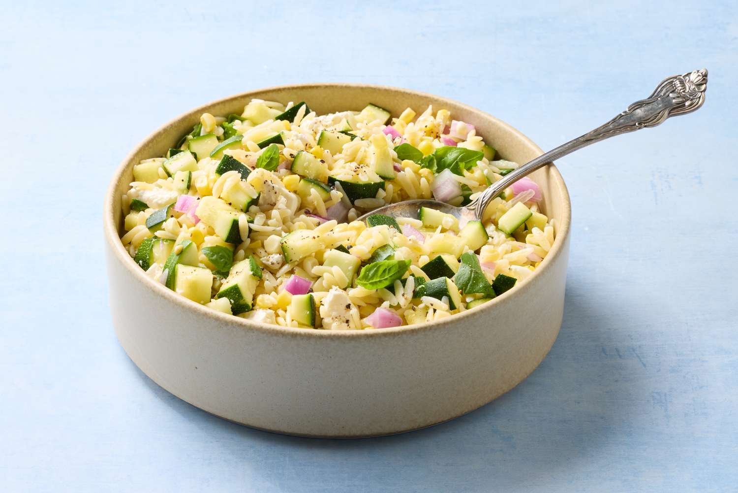 A bowl of corn and zucchini orzo salad topped with basil leaves featuring a serving spoon