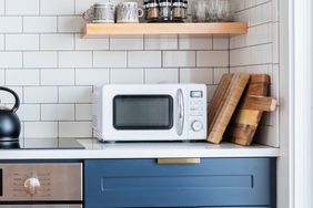 A kitchen counter scene with a microwave wooden cutting boards and a shelf with mugs and jars