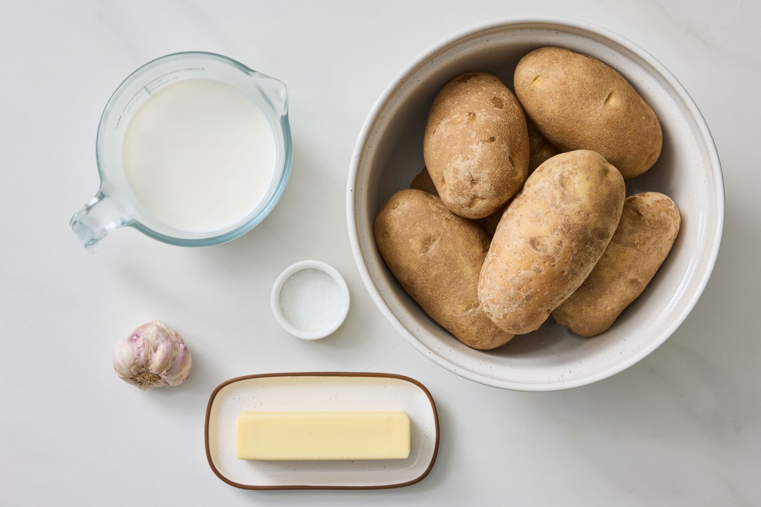 Ingredients for garlic mashed potatoes including potatoes a stick of butter a clove of garlic a measuring cup of milk and a small dish of salt