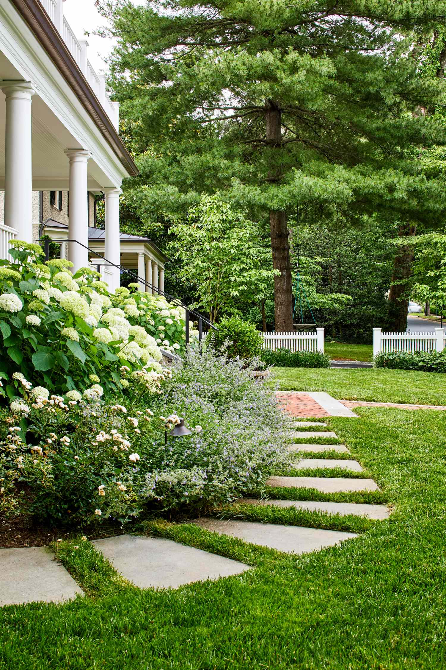 A garden with a path of square stepping stones next to a house and planted flowers with trees in the background
