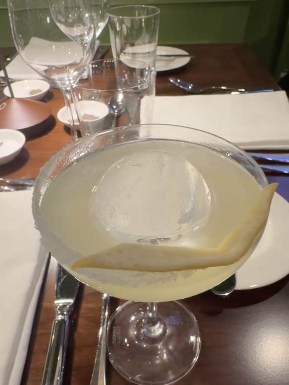 A cocktail served in a glass with a twist garnish placed on a set dining table with napkins and cutlery