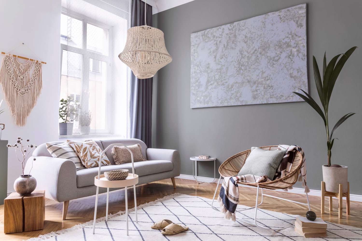 Modern scandi interior design of living room with design rattan armchair, gray sofa, coffee table, beige macrame, plants and elegant accessories.