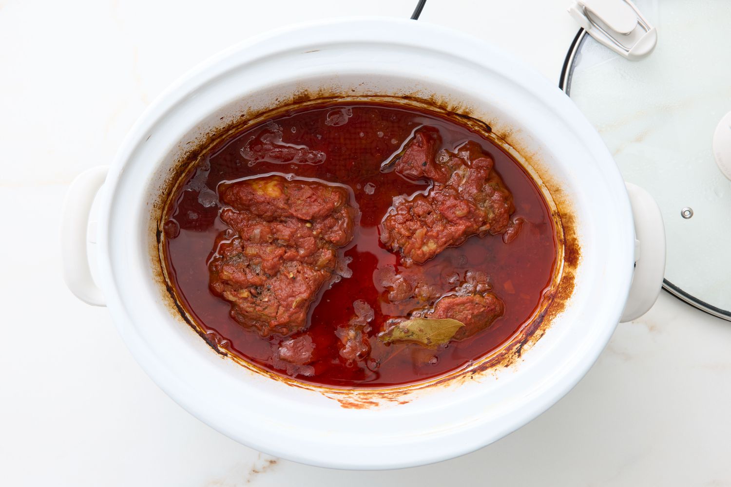 Slow cooker with cooked pork in sauce, lid partially visible