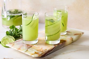 Three cucumber cooler drinks in glasses on a marble tray with lime and mint garnish