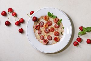 A plate with a pancake topped with cherries and garnished with mint surrounded by whole cherries and mint leaves