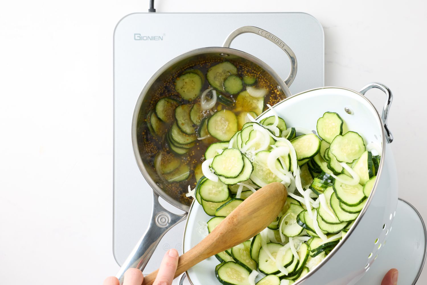 A pot on a stove with sliced cucumbers and onions being poured into it from a colander with a wooden spoon
