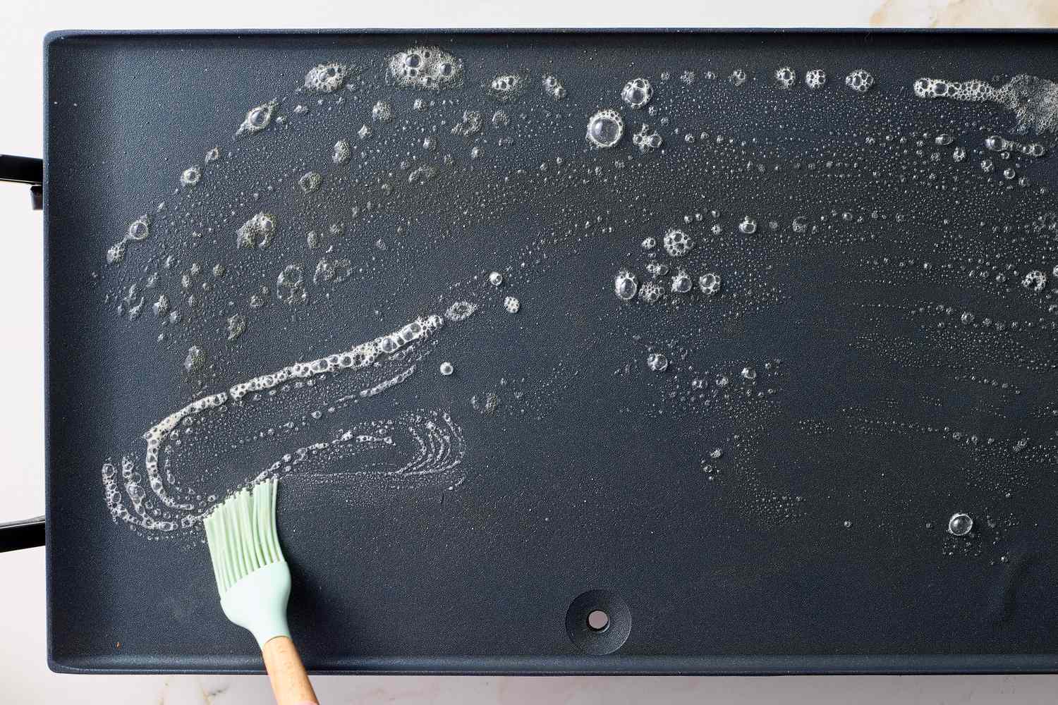 A silicone brush spreading soapy water on a griddle surface