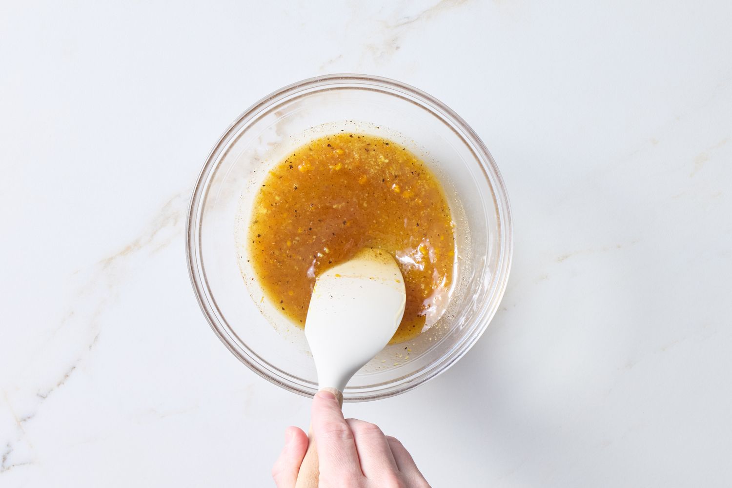A hand stirring honey glaze in a clear glass bowl with a white spatula