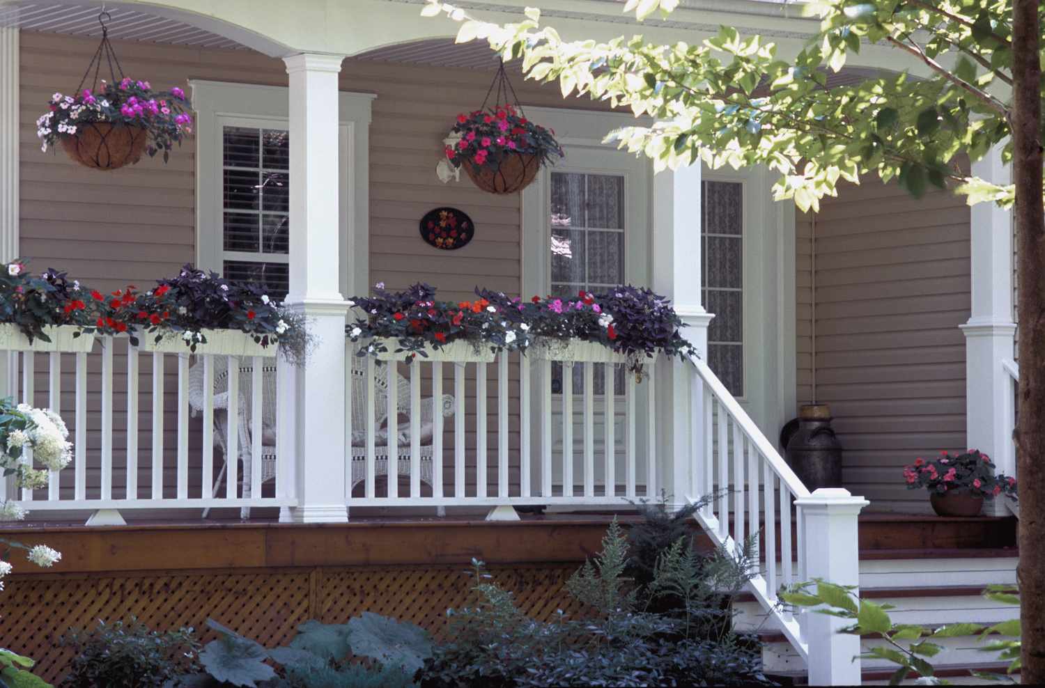 A front porch with hanging and railing flower baskets white railing and a few chairs visible