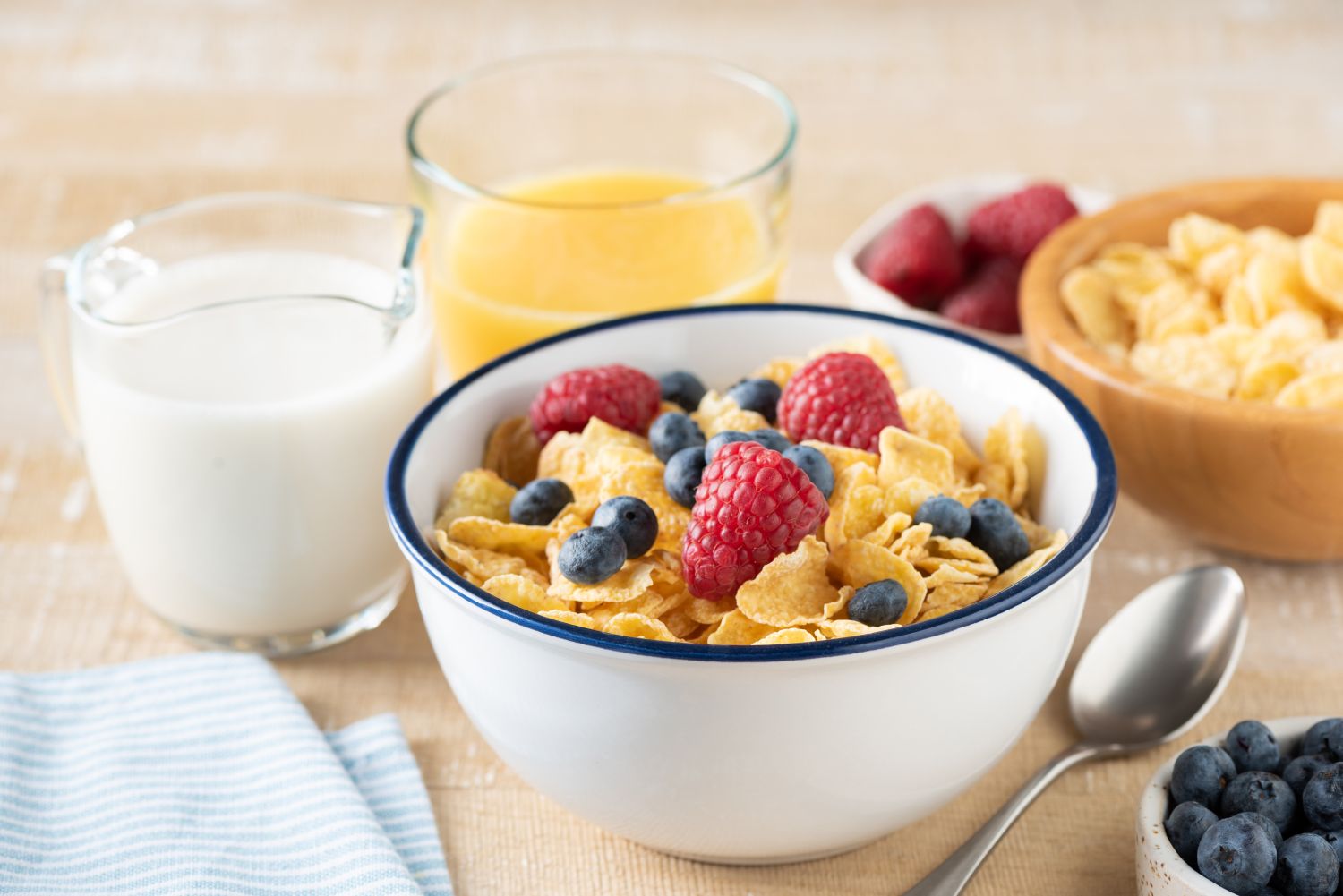 Bowl of cereal topped with raspberries and blueberries surrounded by milk orange juice and bowls of berries and cereal