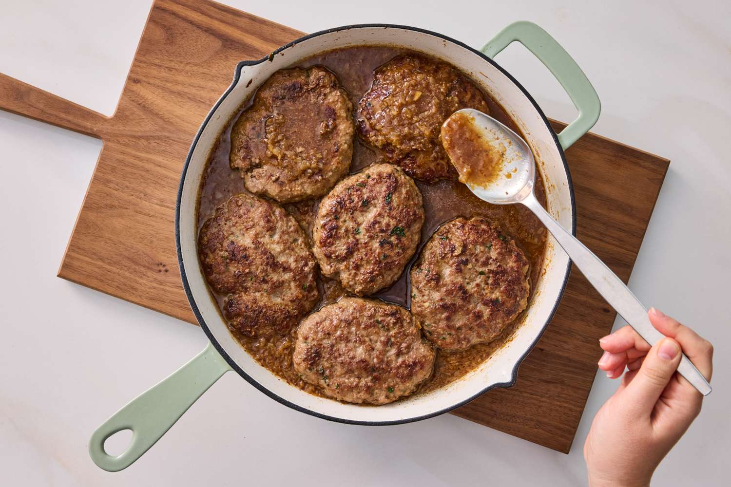 A hand scooping gravy over Salisbury steak patties in a pan