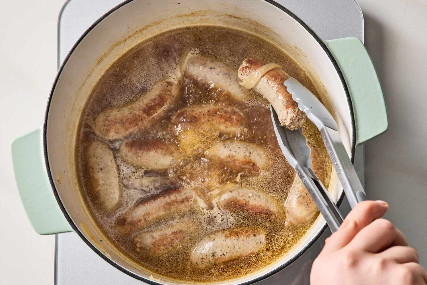 Sausages being cooked in a pot of liquid with a hand using tongs to turn them