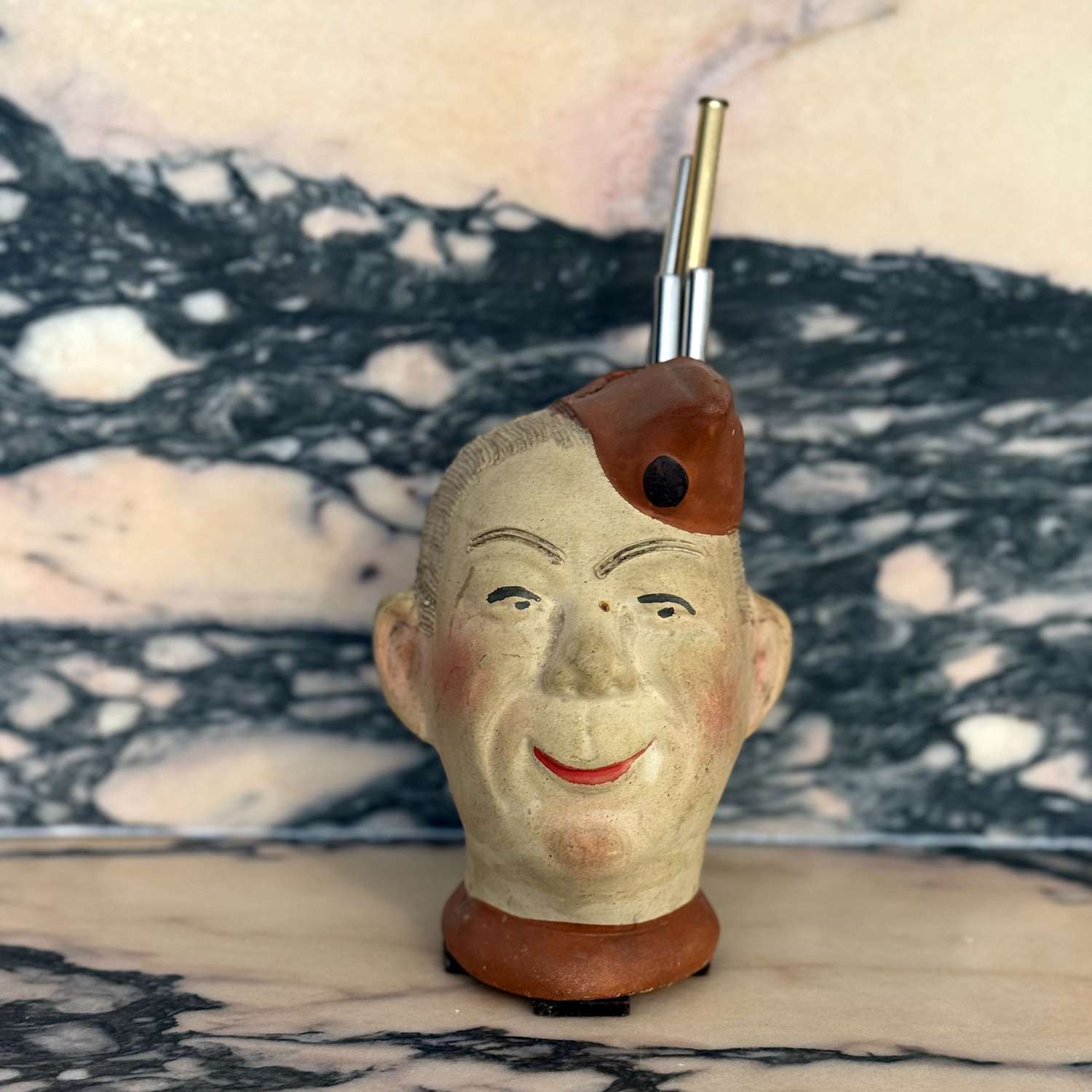 A sculpted head-shaped object with a smiling face and a pipe inserted at the top displayed against a marble background