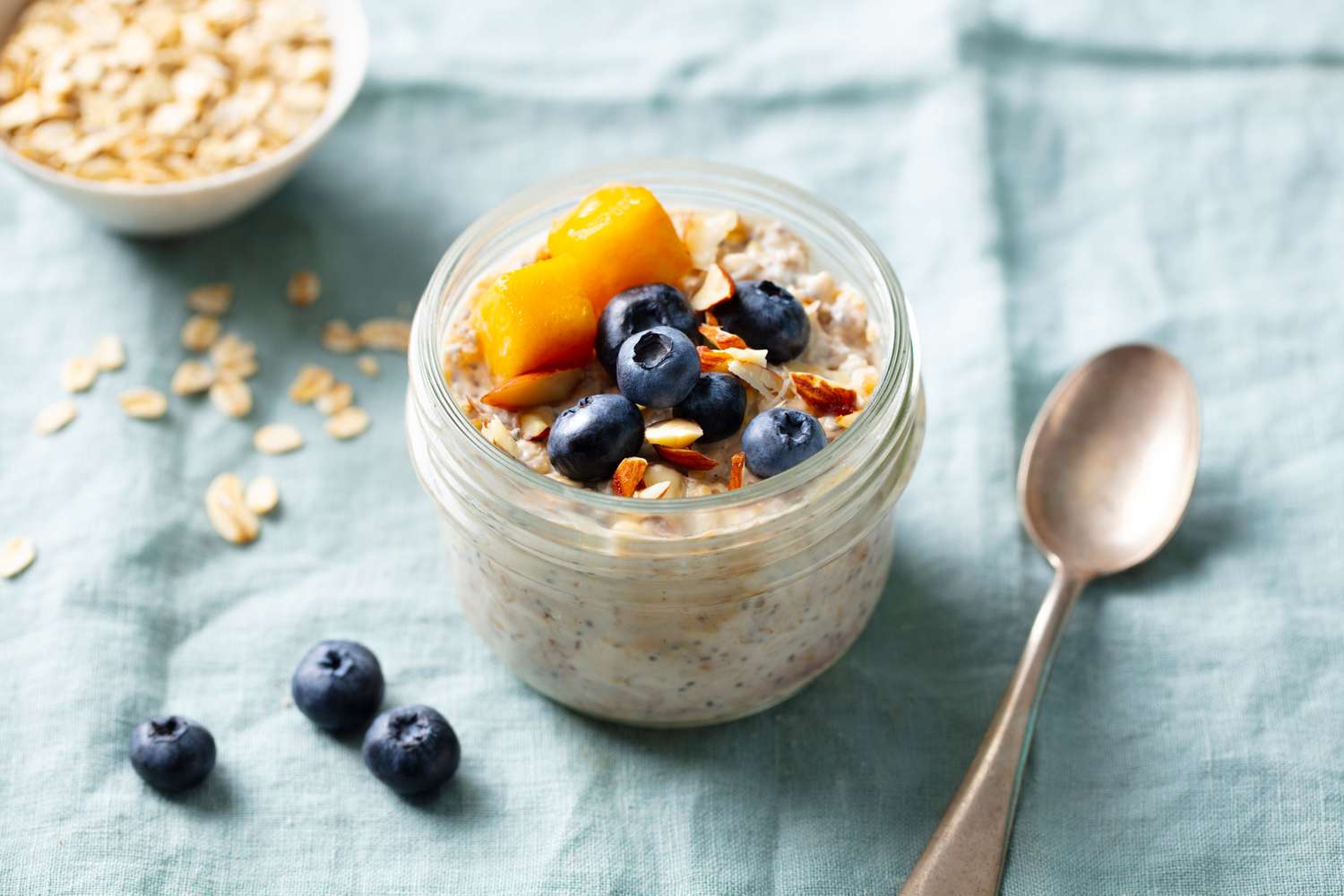 A jar of oatmeal topped with blueberries and mango slices placed on a blue cloth with a spoon nearby and oats scattered around
