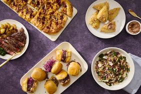 table with purple cloth and varerty of dishes, steak, sliders, grain salad, and more