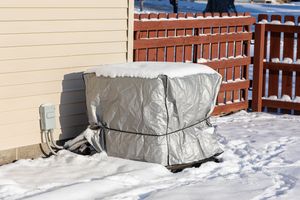 Air conditioning unit covered with a tarp and snow outside a house with a wooden fence in the background